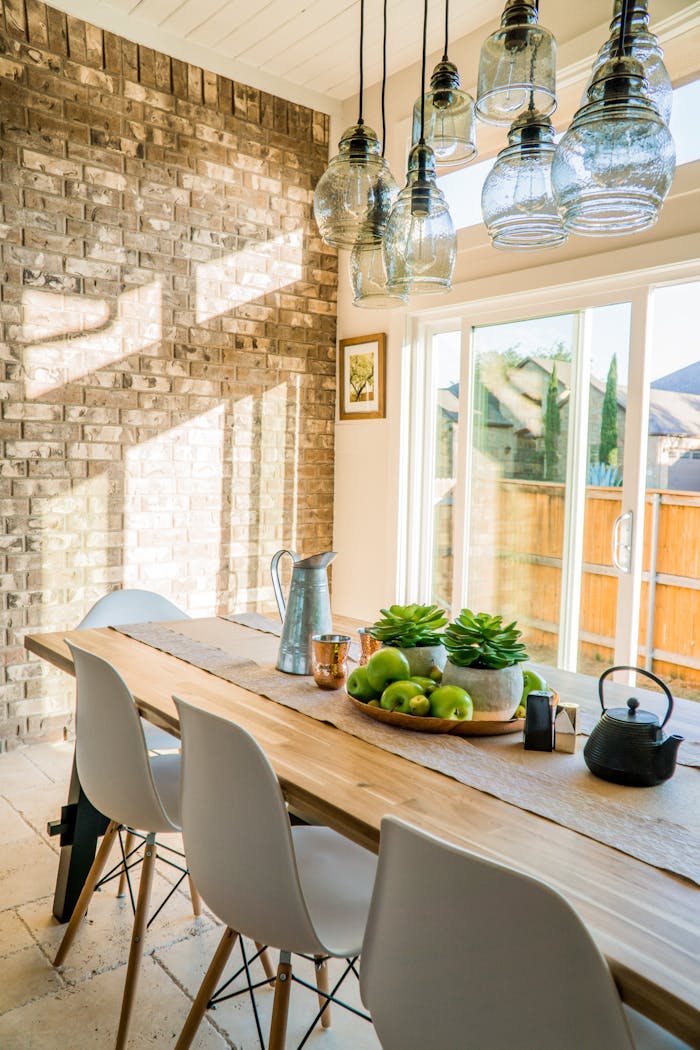 gallery-04 Sunlit dining room with contemporary decor and a rustic brick wall, featuring stylish lighting and a wooden table.